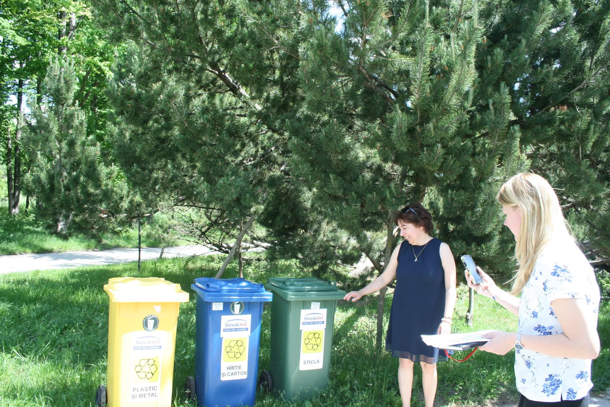 Two women beside yellow, blue and green sorting bins under a large pine tree