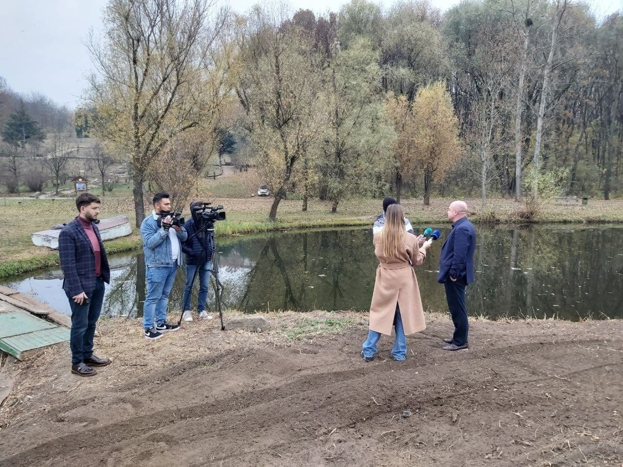 A television interview by a park pond in autumn, with a camera crew present